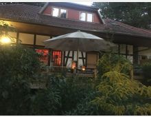 Fachwerkhaus mit rotem Ziegeldach, großer Fensterfront und Holzterrasse, umgeben von dichtem Garten mit gelben Blüten. Vor der Terrasse steht ein Sonnenschirm, und im Inneren des Hauses ist warme Beleuchtung zu sehen.