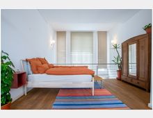 Minimalistisch eingerichtetes Schlafzimmer mit weiß gestrichenen Wänden, einem Holzbett mit orangefarbener Bettwäsche, einem bunten Teppich und einem großen Holzschrank mit Spiegel. Drei große Fenster mit Jalousien und mehrere Zimmerpflanzen ergänzen den Raum.