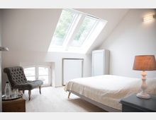 Helles Schlafzimmer mit Dachschrägen, zwei großen Dachfenstern und einer kleinen weißen Wandnische mit Fenster. Im Raum befinden sich ein grauer Polstersessel, ein Bett mit heller Bettdecke, ein kleiner Nachttisch mit Lampe und eine weiße Schranktür.