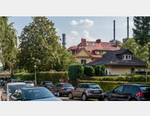 Straßenszene in einem Wohngebiet mit geparkten Autos, dichtem Grün und Einzelhäusern mit roten und schwarzen Dächern im Hintergrund; dahinter ragen Industrie-Schornsteine in den Himmel.