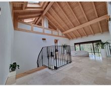 Geräumiger, heller Wohnbereich mit hohen Decken und freiliegender Holzbalkenkonstruktion. Der Raum verfügt über einen offenen Treppenabgang mit schwarzem Geländer, große Fensterfronten mit Blick ins Freie, helle Fliesenböden und dekorative Pflanzen in weißen Töpfen.