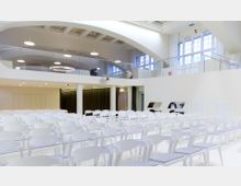 Großer, heller Veranstaltungsraum mit weißen Stühlen in Reihen angeordnet. Der Raum verfügt über hohe Decken mit abgerundeten Bögen, große Fenster mit viel Tageslicht und eine Galerie mit Glasgeländer im oberen Bereich.