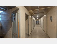 Langer Flur eines alten Gebäudes mit beidseitig angeordneten, kleinen Zellen mit Türen, die teils geöffnet sind. Die Wände sind mit weißen Fliesen verkleidet, und im Hintergrund ist eine geschlossene Tür mit Fenstereinsatz sichtbar.