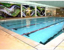 Hallenbad mit einem großen rechteckigen Schwimmbecken, das durch Leinen in Bahnen unterteilt ist. An der Wand befindet sich ein buntes Mosaik mit wellenförmigen Mustern, und im Hintergrund sind Sitzgelegenheiten sowie Fenster und Türen zu sehen.