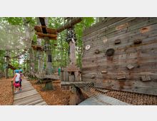 Kletterpark im Wald mit Plattformen, Seilbrücken und einem Kletterelement aus Holz. Im Vordergrund ein hölzerner Weg, zwei Personen mit Helmen stehen darauf, umgeben von Bäumen mit befestigten Kletterstrukturen.