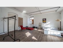 Ein einfacher, heller Raum mit Holzfußboden, weißen Wänden und hohen Decken. Der Raum enthält wenige Möbelstücke, darunter ein roter Sessel, eine rote Liege, einen Kleiderständer sowie vereinzelte Dekorationen wie ein Poster und einen runden Spiegel vor einem Fenster.