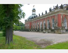 Historisches Gebäude mit roter Backsteinfassade, großen Rundbogenfenstern und steinernen Skulpturen auf Balustraden. Im Hintergrund ist eine Kuppel mit vergoldetem Wetterhahn zu sehen, umgeben von einem Park mit Bäumen und einer Freifläche.