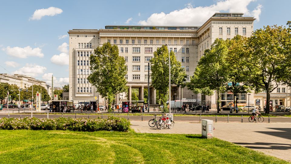 Großes, helles Stein-Gebäude im Stil der frühen Moderne mit vielen Fenstern und einem hohen Durchgang im Erdgeschoss, davor eine breite Straße mit Haltestellen und Bäumen. Im Vordergrund liegt eine Rasenfläche mit Blumenbeet und Radweg, umgeben von städtischer Bebauung.