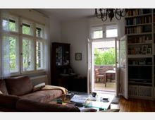 Wohnzimmer mit großen Sprossenfenstern und einer offenen Terrassentür, die auf eine Backstein-Terrasse mit Tisch und Stühlen führt. Der Raum enthält eine braune Polstercouch, einen Couchtisch aus Glas, eine Bücherwand und einen Kronleuchter, mit viel Tageslicht, das durch die Fenster fällt.