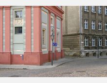 Ecke einer historischen Straße mit einem hellrosa und grauen Gebäude mit klassizistischen Säulen und Fenstern; daneben ein älteres Gebäude mit beigem Putz und weißen Fensterrahmen. Steinpflaster bedeckt die Straße, und ein Laternenpfahl sowie ein Verkehrsschild sind sichtbar.