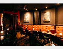 Dunkel beleuchteter Bar- oder Lounge-Innenraum mit langen, orangefarbenen, gepolsterten Sitzbänken entlang der Wand, kleinen runden Tischen mit Lampen und schwarzen Stühlen. An den dunkelgrauen Wänden hängen gerahmte Bilder, und schwere Vorhänge sowie Deckenstrahler prägen die Atmosphäre.