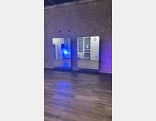 Dance or fitness studio with a wooden floor and an exposed brick wall, featuring two large freestanding mirrors on wheeled stands. Reflections show tall windows with blinds, a radiator, and studio equipment, with blue lighting cast across the room.