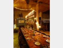 Ein festlich gedeckter Holztisch in einem rustikalen Raum mit Ziegel- und Holzwänden; darüber ein Kronleuchter aus Kerzen und Blumenarrangements. Der Raum ist mit warmem Licht beleuchtet und wirkt wie eine umgebaute Scheune mit klassischen landwirtschaftlichen Elementen im Hintergrund.