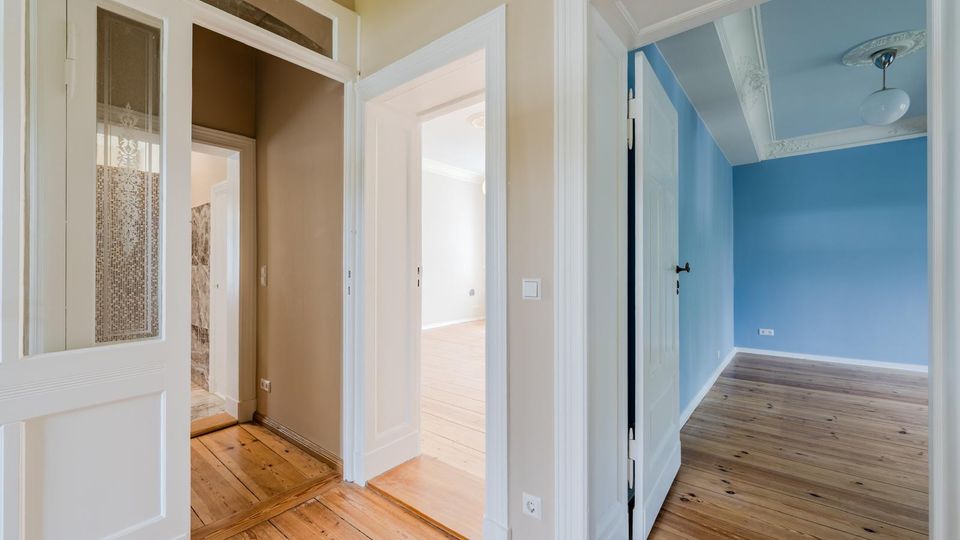 Innenansicht eines Wohnungsflurs mit hellen Holzdielen, hohen weißen Türzargen und mehreren offenen Durchgängen zu angrenzenden Räumen. Rechts ist ein blau gestrichenes Zimmer mit Stuck an der Decke und Deckenleuchte sichtbar, links eine Tür mit Glasfeld und dekorativem Muster.