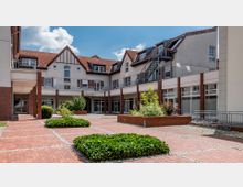 Innenhof eines modernen Gebäudekomplexes mit roten Ziegeldächern und weißer Fassade, umgeben von hohen Fenstern und Balkonen. Der Boden ist mit roten Pflastersteinen bedeckt, und in der Mitte befinden sich bepflanzte Beete mit grüner Vegetation.