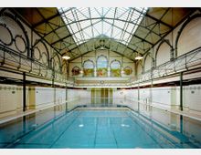 Historisches Hallenbad mit großer Schwimmhalle, gekachelten Wänden und verzierten Bögen. Die Decke besteht aus einer Stahlkonstruktion mit Glasoberlichtern, und an der Stirnwand befinden sich bunte Wandmalereien.
