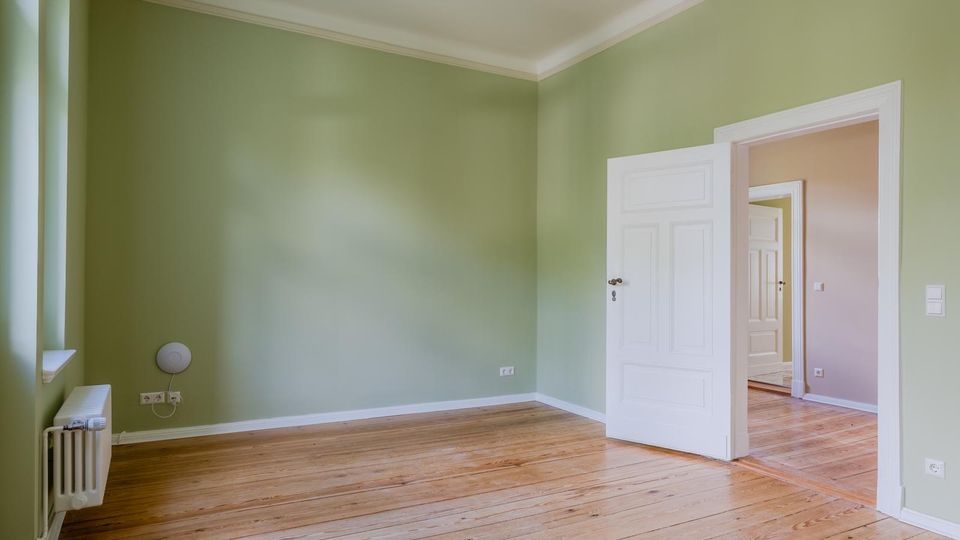 Leerer Innenraum mit hellgrün gestrichenen Wänden, hohen Decken mit weißer Stuckleiste und breiten Holzdielen. Links ein Fenster mit Heizkörper darunter, rechts eine weiße Kassettentür, die in einen weiteren Raum mit ähnlichem Holzboden führt.