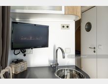 Kleine Küchenzeile in modernem Design mit einer Edelstahlspüle, einem wandmontierten Flachbildfernseher und einer hellen Tür mit Bullaugenfenster. Im Hintergrund sind ein Regal und ein Teil eines Raumes sichtbar.