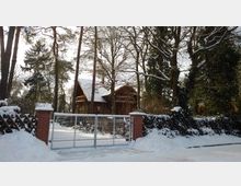 Schneebedecktes Grundstück mit einem großen Holzhaus im Hintergrund, umgeben von hohen Bäumen. Im Vordergrund ein verschlossenes Metalltor mit roten Backsteinpfeilern.