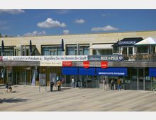 Moderne Gebäudeansicht in Potsdam mit Glasfassade im Erdgeschoss, über der sich gastronomische Einrichtungen mit Sonnenschirmen und Sitzbereichen befinden. Im Vordergrund sind Schilder der „Weisse Flotte Potsdam“ sowie Personen auf einer gepflasterten Fläche zu sehen.