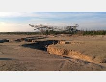 Weite Sandfläche mit erodierten Gräben im Vordergrund und einer großen Industriekonstruktion im Hintergrund, vermutlich ein Förderband oder Abbaugerät, umgeben von einem dünnen Baumstreifen unter einem hellen Himmel.