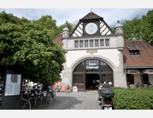 Eingang des Bahnhofs Berlin-Grunewald mit historischer Fassade im Fachwerkstil, einer großen Uhr im Giebel und einem Bogenfenster mit dem Stationsnamen. Im Vordergrund stehen Fahrräder und ein Baum, rechts ist ein Café zu sehen.