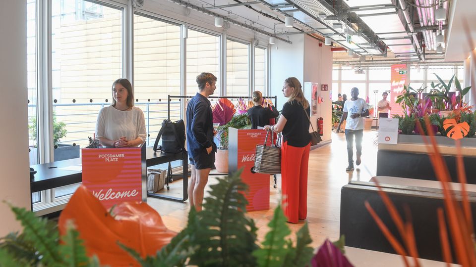 Bright modern office lobby with large floor-to-ceiling windows, exposed ceiling pipes and cable trays, and light wood flooring. Welcome check-in counters and coat racks sit among indoor plants and lounge seating along a wide corridor.