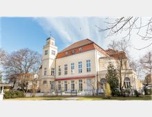 Gelbes, historisches Gebäude mit einem kleinen Turm, großen Fenstern und einem roten Ziegeldach, umgeben von kahlen Bäumen und einem gepflegten Vorgarten unter klarem Himmel.