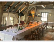 Rustikal eingerichteter Veranstaltungsraum mit Holzbalkendecke, großen Fenstern und langen Tafeln mit weißer Tischdecke, gedeckt für ein Essen mit Kaffeetassen, Karaffen, Blumenarrangements und Hängelampen zur Beleuchtung.