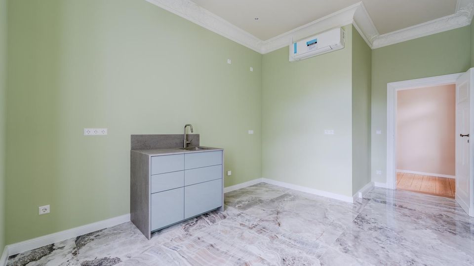 Heller, leerer Innenraum mit hellgrünen Wänden, weißer Stuckdecke und glänzendem Marmorfußboden. An der Wand steht ein kleiner Unterschrank mit Spüle; darüber ist eine Klimaanlage montiert, rechts führt eine Tür in einen angrenzenden Raum.