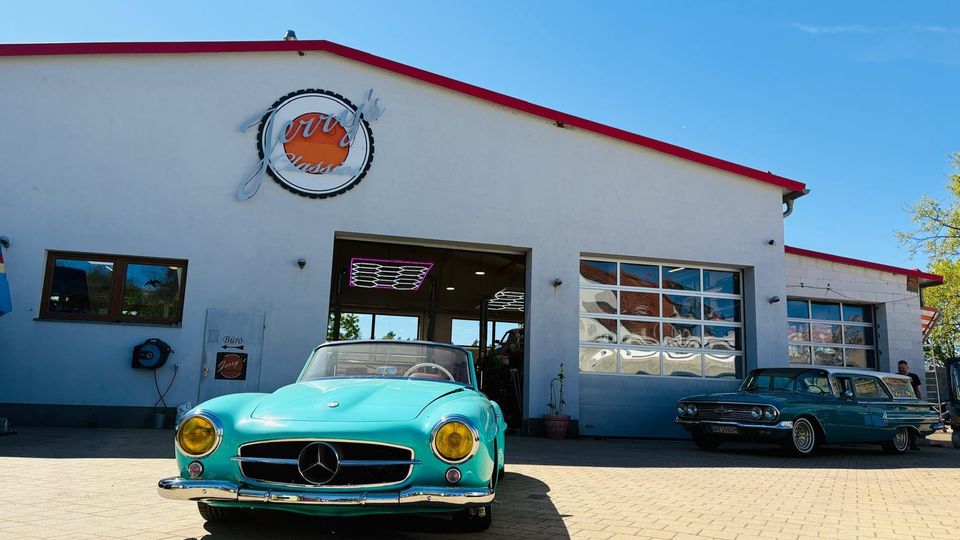 Frontansicht einer Autowerkstatt mit weißer Fassade und rotem Dachrand, großem Einfahrtstor und einem Fenster‑Rolltor an der Seite. Davor liegt ein gepflasterter Hof, auf dem Oldtimer-Fahrzeuge vor dem geöffneten Werkstatteingang stehen.
