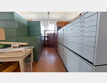 Schmaler Raum mit hohen Metallschränken auf beiden Seiten, ausgestattet mit zahlreichen flachen Schubladen in grüner und weißer Farbe. Im Hintergrund befindet sich eine kleine Holzleiter vor einer Fenstertür, und ein Holztisch links trägt gerollte und gestapelte Papiere.