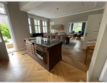 Wohnzimmer mit offener Küche und Holzboden im Fischgrätmuster. Ein zentraler Küchenblock mit Kochfeld steht vor einem Essbereich mit großem Tisch und Stühlen, im Hintergrund ein weiterer Sitzbereich mit Sofas und Sesseln, umgeben von großen Fenstern mit Blick ins Grüne.