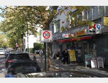 Straßenszene mit parkenden Autos, einem Baum und einem Verkehrsschild für eine Tempo-30-Zone. Rechts sind Geschäfte mit Werbeschildern und Zeitungsständern unter einem Gebäudevordach zu sehen.