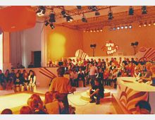 Ein Fernsehstudio mit Publikum auf Sitzrängen und einer Bühne, auf der Schriftzug „Are u hot?“ zu sehen ist. Im Vordergrund befindet sich ein Kameramann, und im Studio sind Beleuchtungseinrichtungen an der Decke befestigt.