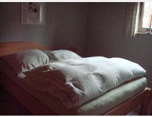 Ein einfach eingerichtetes Schlafzimmer mit einem hölzernen Doppelbett, das mit weißen, gestreiften Bettdecken bezogen ist. Im Hintergrund sind ein kleines Fenster mit einer Gardine und ein gerahmtes Bild an der Wand zu sehen.