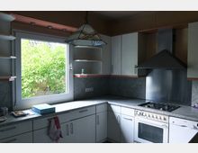 Ein heller Küchenraum mit einer L-förmigen Arbeitsplatte aus grauem Stein, hellen Holzmöbeln und einem großen Fenster mit Blick auf grüne Büsche. Links vom Fenster befinden sich offene Regale, rechts eine Dunstabzugshaube aus Edelstahl über einem Gasherd.