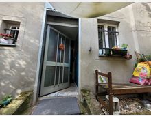 Eingangsbereich eines Wohnhauses mit geöffneter Glastür, flankiert von zwei Fenstern mit Blumenkästen und Gitterstäben. Im Vordergrund stehen eine Holzbank, Gartenzubehör und Säcke mit Blumenerde auf einem Kiesboden.