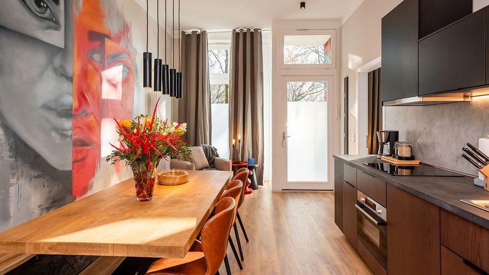 Moderner, schmaler Küchen- und Essbereich mit langem Holztisch, braunen Stühlen und schwarzen Pendelleuchten sowie einer Küchenzeile mit dunklen Schränken und grauer Arbeitsplatte. An der linken Wand befindet sich ein großformatiges Wandgemälde, am Ende des Raums führen hohe Fenster und eine Glastür mit Vorhängen nach außen.