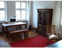 Ein heller Raum mit Holzboden, ausgestattet mit einem rustikalen Holztisch und passender Bank, einem Holzschrank mit Glastüren, einem Holzstuhl mit Kissen und einem roten Teppich. Große Fenster mit weißen Vorhängen lassen Tageslicht herein, und auf dem Tisch stehen ein Computerbildschirm, eine Schreibtischlampe und Dekorationsgegenstände.