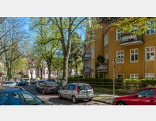 Straßenszene mit gelbem Wohngebäude auf der rechten Seite, mehreren Balkons, und Bäumen, die einen Schatten werfen. Im Vordergrund parken Autos entlang einer Kopfsteinpflasterstraße, während ein Straßenschild mit der Aufschrift „Düppelstraße“ sichtbar ist.