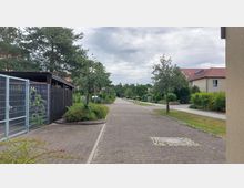 Eine ruhige Wohnstraße mit gepflastertem Weg, gesäumt von kleinen Bäumen und Sträuchern. Rechts stehen moderne Einfamilienhäuser mit roten Ziegeldächern, während links eine offene Metallzaunanlage und ein Carport sichtbar sind.