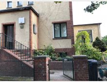 Gelbes Wohnhaus mit Backsteinrahmen um Fenster und Türen, einer Treppe mit Metallgeländer, einem kleinen Vorgarten mit Pflanzen und einem Tor aus Metall und Backstein. Im Hintergrund ist ein weiteres Gebäudeteil sowie Sträucher sichtbar.