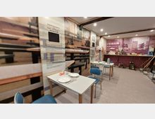 Innenraum einer musealen Ausstellung mit Sitzbereich, bestehend aus einfachen Tischen und blauen Stühlen. An den Wänden befinden sich großformatige Fotos und Informationstafeln über die Arbeiterverpflegung im Stahl- und Walzwerk Brandenburg; im Hintergrund wird eine historische Kantinen- und Verkaufsumgebung mit Alltagsgegenständen dargestellt.