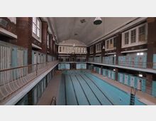 Innenraum eines historischen Schwimmbads mit leeren Schwimmbecken, umgeben von zwei Etagen mit Umkleidekabinen in hellblauer Farbe. Die roten Backsteinwände, hohen Fenster und gewölbte Decke verleihen dem Raum einen architektonisch klassischen Charakter.
