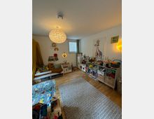 Kinderzimmer mit Holzboden, einer Spielmatte und einem hellgrauen Teppich. An den Wänden befinden sich Regale mit Spielzeug und Büchern, eine Giraffen-Wanddekoration, ein Bett mit gelbem Vorhang sowie eine Hängelampe mit runder, strukturierter Leuchte. Ein kleines Fenster lässt Tageslicht herein.