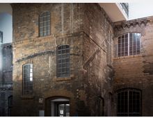 Backstein-Gebäude mit hohen, schmalen Bogenfenstern und deutlichen Abnutzungsspuren an der Fassade; im unteren Bereich ein Eingang mit Glastür und Hinweiszeichen.