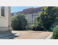 Eine asphaltierte Fläche mit gepflastertem Rand führt durch eine kleine grüne Parkanlage mit einfassenden Bäumen. Im Hintergrund ist ein mehrstöckiges Gebäude mit rotem Dach und großen Fenstern zu sehen, links ein Teil eines historischen Gebäudes mit hellen Fassaden.