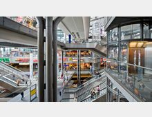 Innenaufnahme eines modernen Bahnhofs mit mehreren Etagen, breiten Rolltreppen, gläsernen Aufzügen und Geschäften wie McDonald's, Burger King und Kaiser's. Große Metallstützen und Glasflächen dominieren die Architektur, während Menschen durch die Ebenen des Gebäudes gehen.