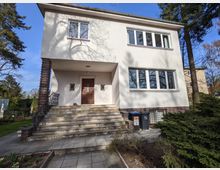 Zweistöckiges, weiß verputztes Wohnhaus mit flachem Dach und großen Fensterfronten, der Eingang liegt erhöht unter einem überdachten Vorsprung. Breite Steintreppe mit Geländer führt zur Holztür; davor gepflasterter Weg in einem Garten mit Bäumen und Sträuchern.